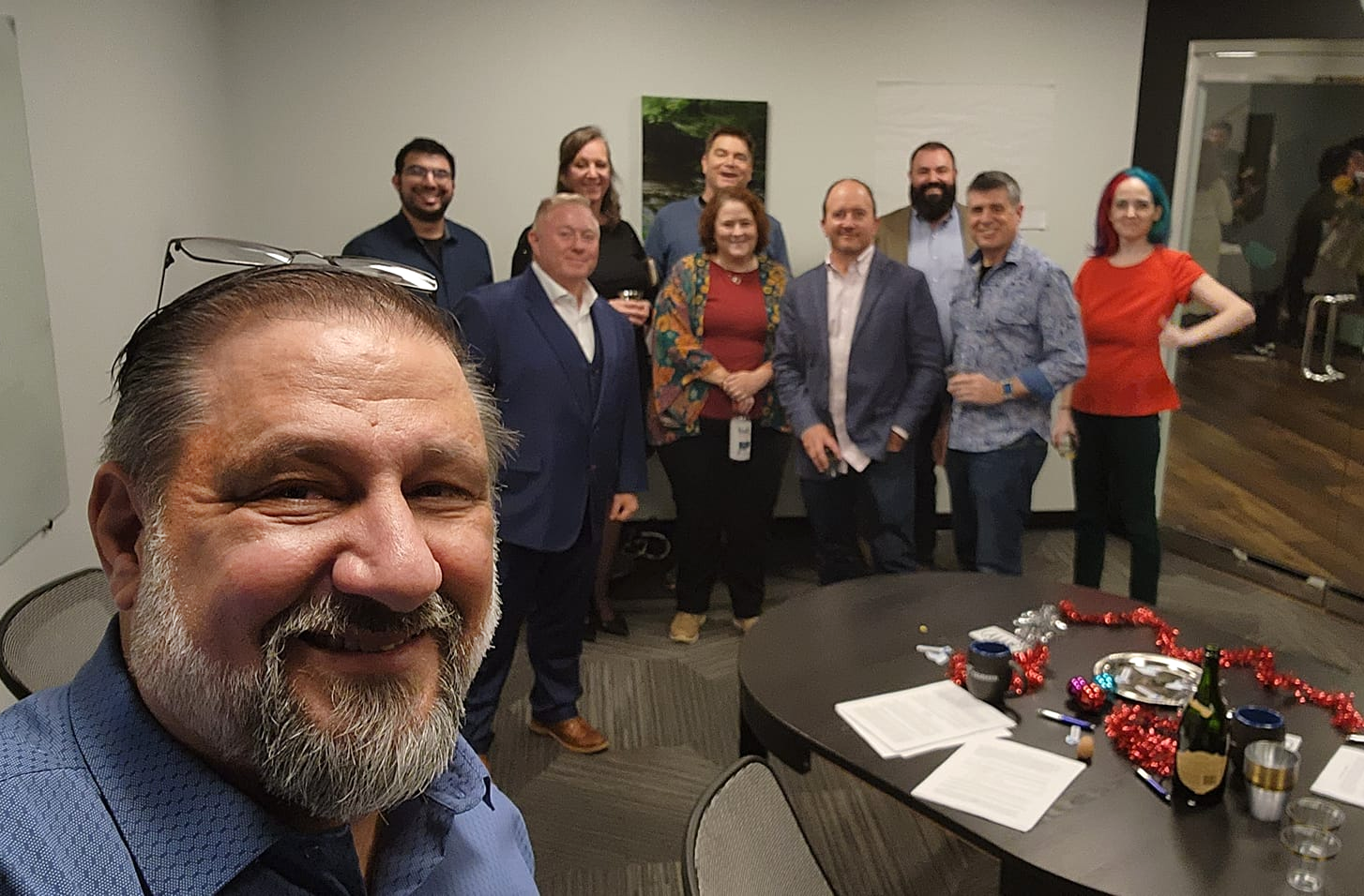 Dax takes a group photo at the signing party for the merger with Chad Chelius, Jeff Tamburino and several other members of the Tamman team. The merger documents and pens are on the table in the foreground, along with celebratory champagne.
